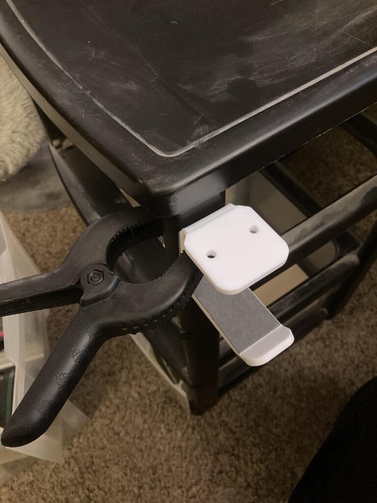 Plastic under-desk or on-wall headphone hanging bracket sticky taped to a plastic drawer cabinet and held on with a plastic clamp.